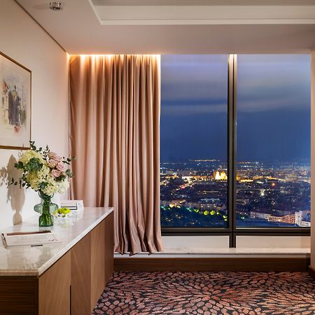 Grand Hotel Millennium صوفيا المظهر الخارجي الصورة The photo depicts a modern hotel room or a lounge area with a stunning view of a cityscape at night. On the left, there's a wooden console table with a vase of flowers and a bottle of water. A piece of art is framed on the wall above the table. The r
