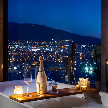 Grand Hotel Millennium صوفيا المظهر الخارجي الصورة The photo features a luxurious bathtub placed by a large window, showcasing a scenic city view at night. There is a wooden tray resting on the edge of the tub, displaying a bottle of champagne, two glass flutes, and several lit candles, creating an a