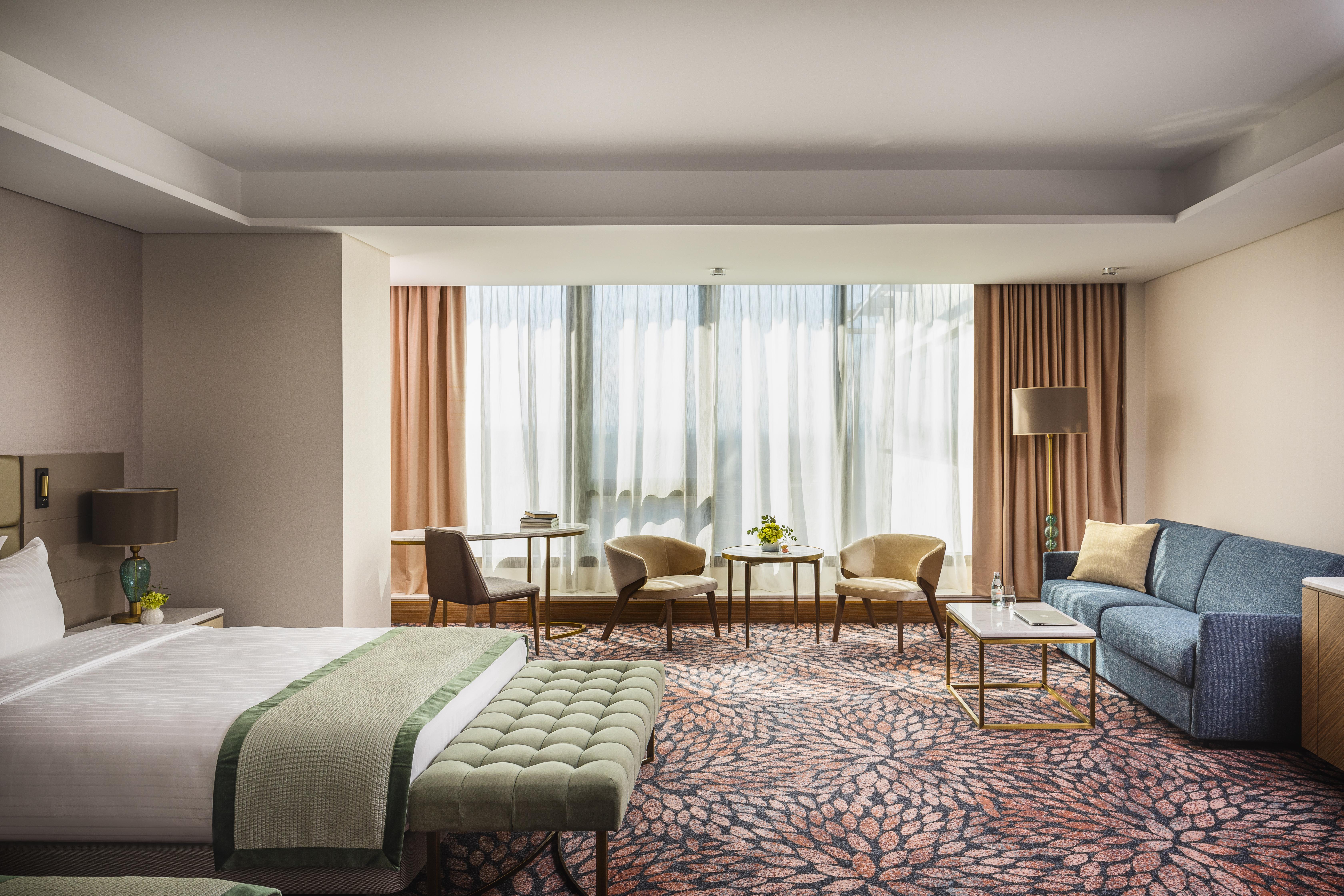 Grand Hotel Millennium صوفيا المظهر الخارجي الصورة The photo shows a modern hotel room featuring a spacious layout. On one side, there is a large bed with a green throw blanket and a tufted bench at its foot. The room is well-lit by large windows dressed with light curtains, which allow natural light