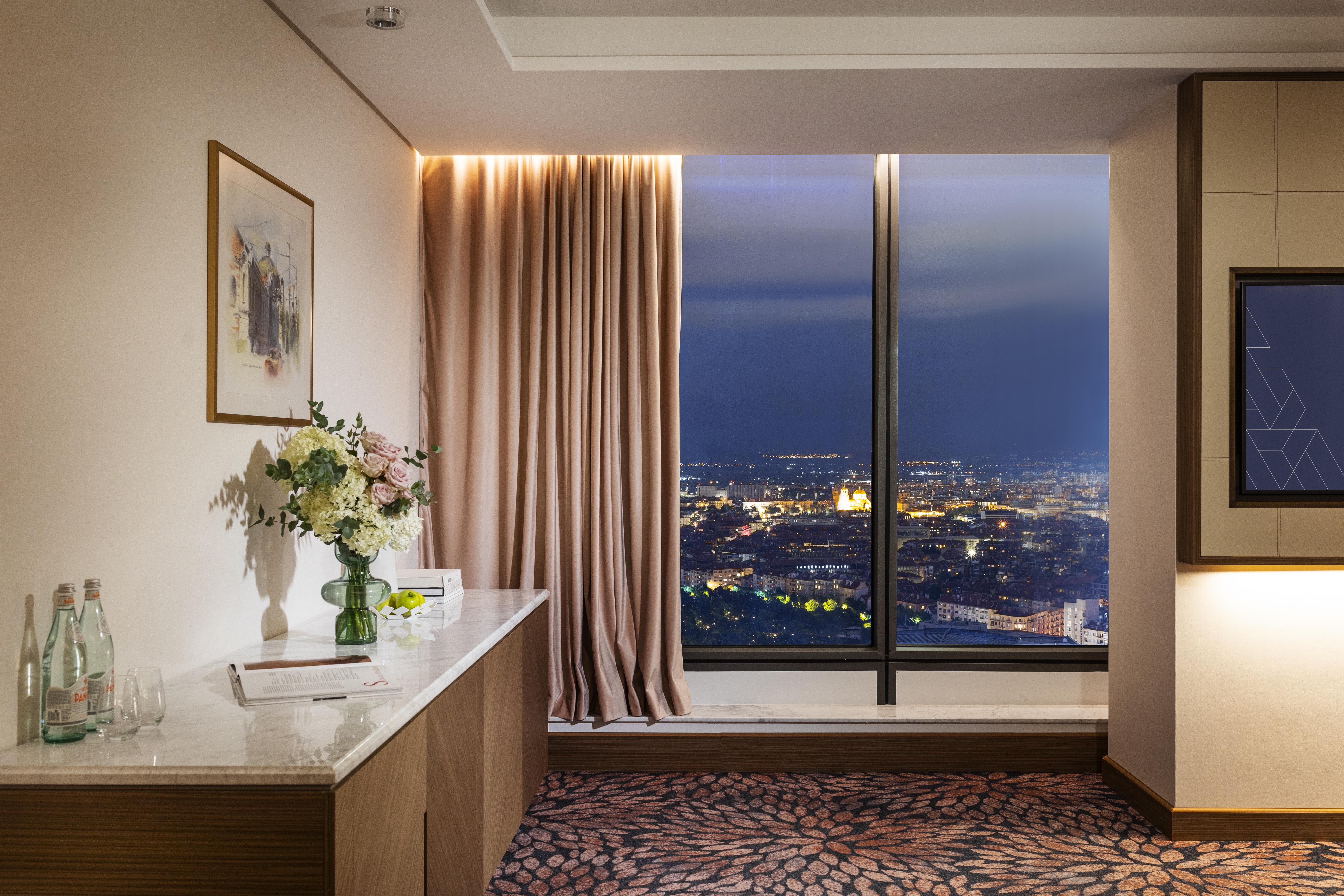 Grand Hotel Millennium صوفيا المظهر الخارجي الصورة The photo depicts a modern hotel room or a lounge area with a stunning view of a cityscape at night. On the left, there's a wooden console table with a vase of flowers and a bottle of water. A piece of art is framed on the wall above the table. The r