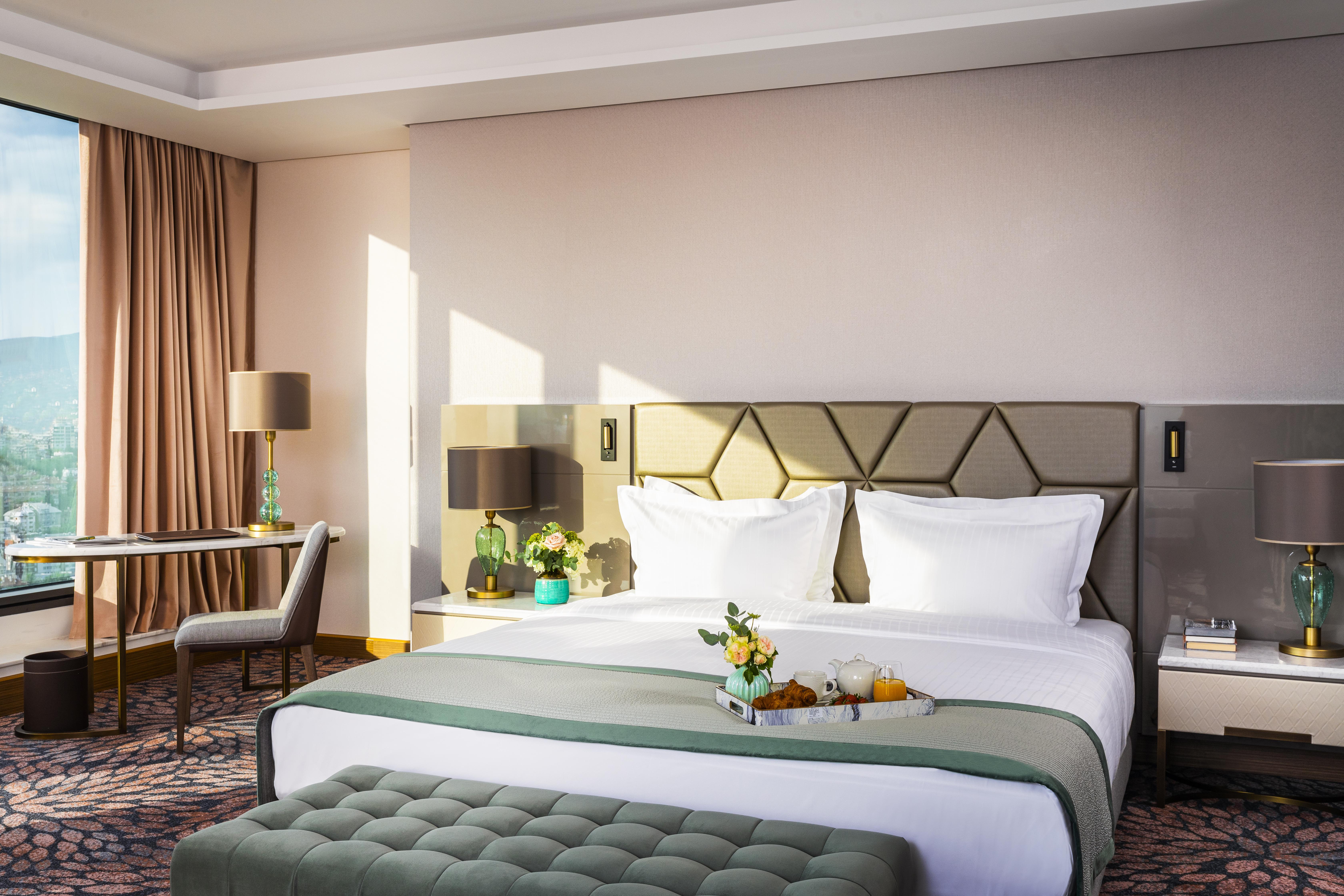 Grand Hotel Millennium صوفيا المظهر الخارجي الصورة The photo depicts a stylish hotel room featuring a large bed with white linens and a green decorative throw. Behind the bed is a geometric-patterned headboard. To the side, there are modern bedside lamps and a small table with a vase of flowers. A se