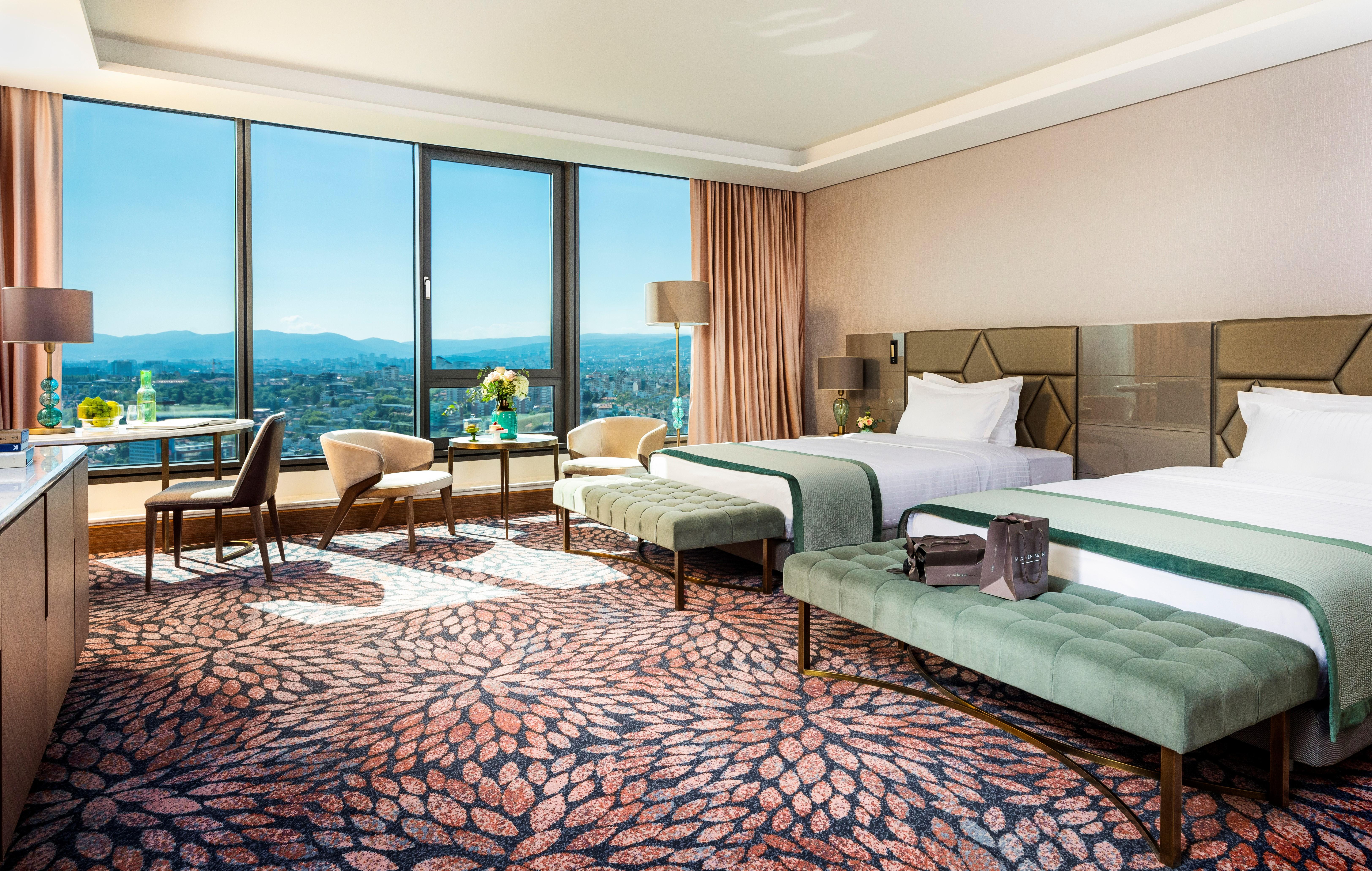 Grand Hotel Millennium صوفيا المظهر الخارجي الصورة The photo depicts a luxurious hotel room featuring two beds with elegant bedding. The room has large windows that offer a panoramic view of the city and mountains in the background. There is a decorative area rug with a floral pattern covering the fl