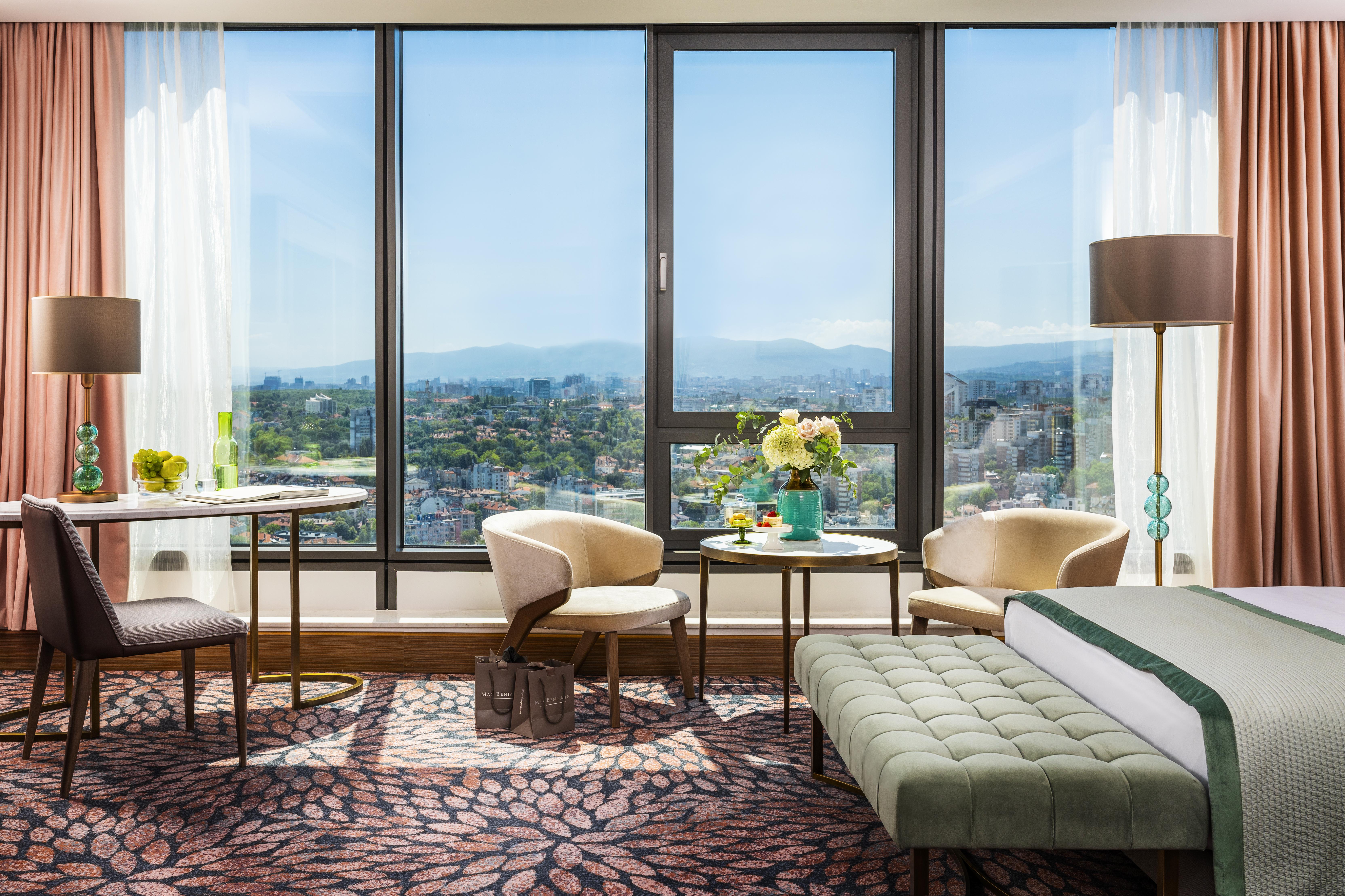Grand Hotel Millennium صوفيا المظهر الخارجي الصورة The photo shows a modern and well-designed hotel room featuring a large window that offers a view of the outdoors, including mountains in the background. Inside the room, there is a neatly made bed with a green accent blanket, a stylish rug with a fl