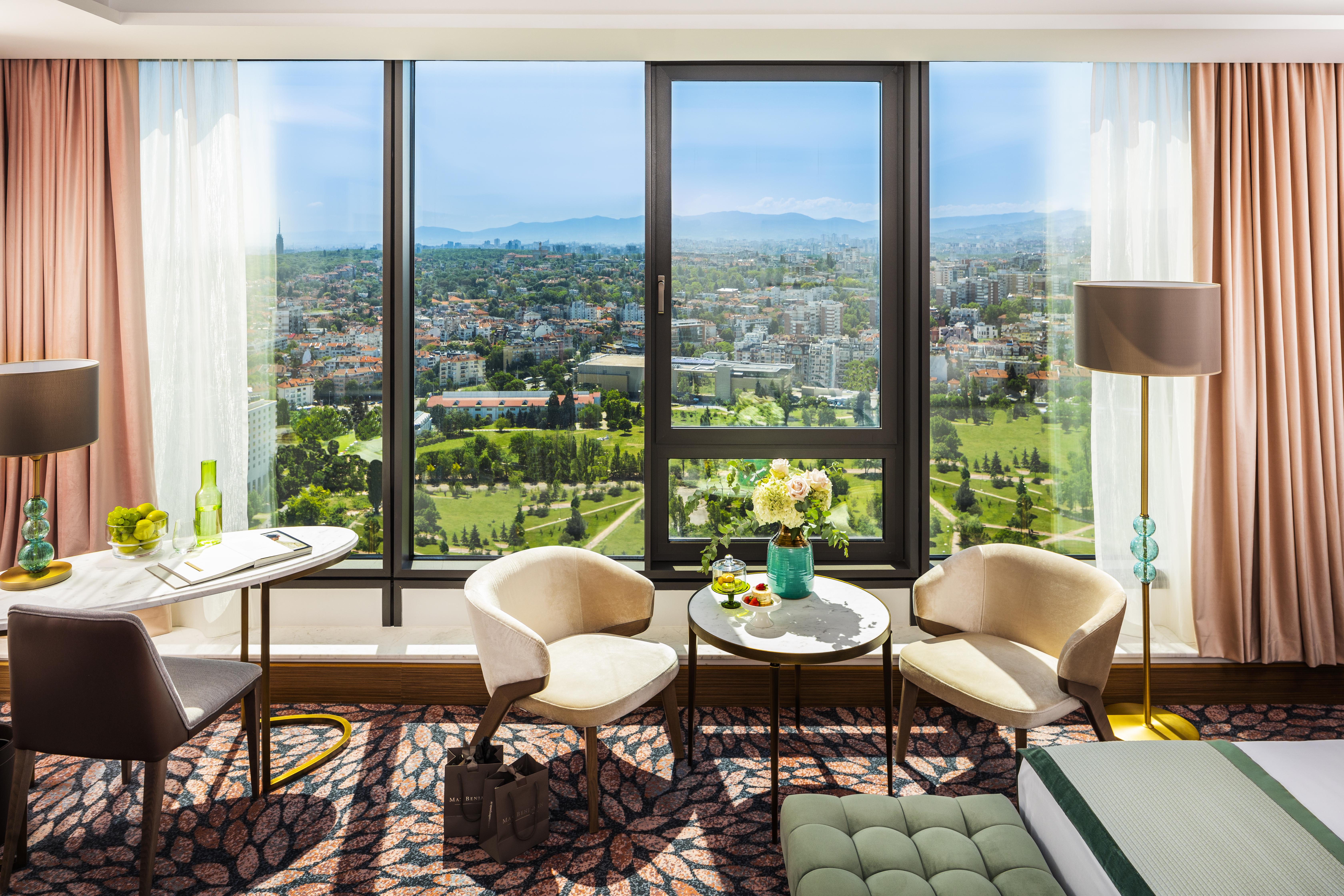Grand Hotel Millennium صوفيا المظهر الخارجي الصورة The photo shows a stylish hotel room with large windows providing a beautiful view of a cityscape and landscape beyond. There are two modern chairs and a small round table set near the windows, accompanied by a vase of fresh flowers. The room feature