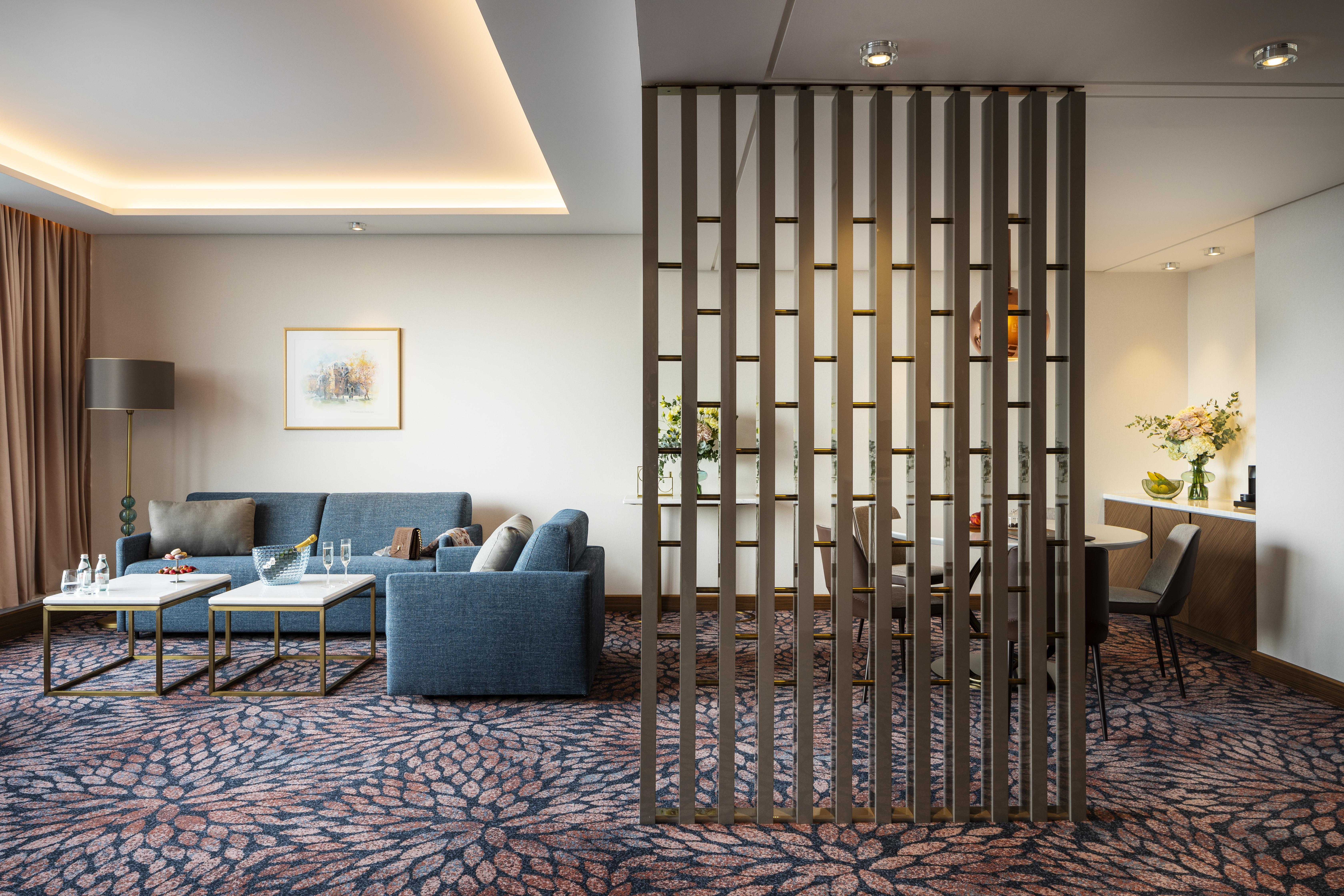 Grand Hotel Millennium صوفيا المظهر الخارجي الصورة The photo shows a modern living space with a comfortable seating area featuring a blue couch and two coffee tables. There is a decorative partition made of vertical wooden slats, which separates this sitting area from a dining space. The dining area 