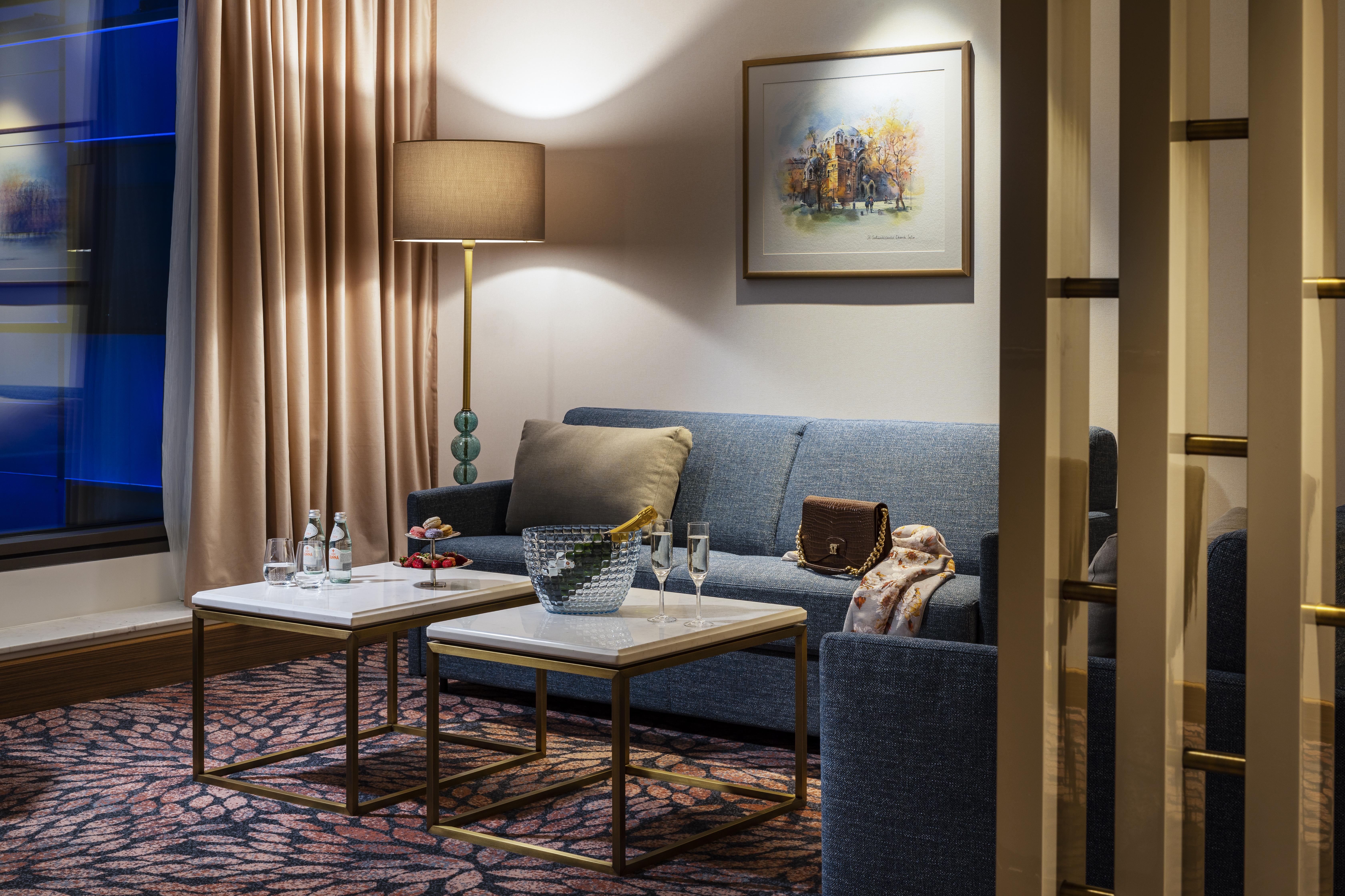 Grand Hotel Millennium صوفيا المظهر الخارجي الصورة The photo shows a cozy and elegantly decorated living space. It features a blue sofa against a wall, complemented by a painting above it. There are two small marble-top coffee tables with gold frames, one of which holds decorative items and glasses. 