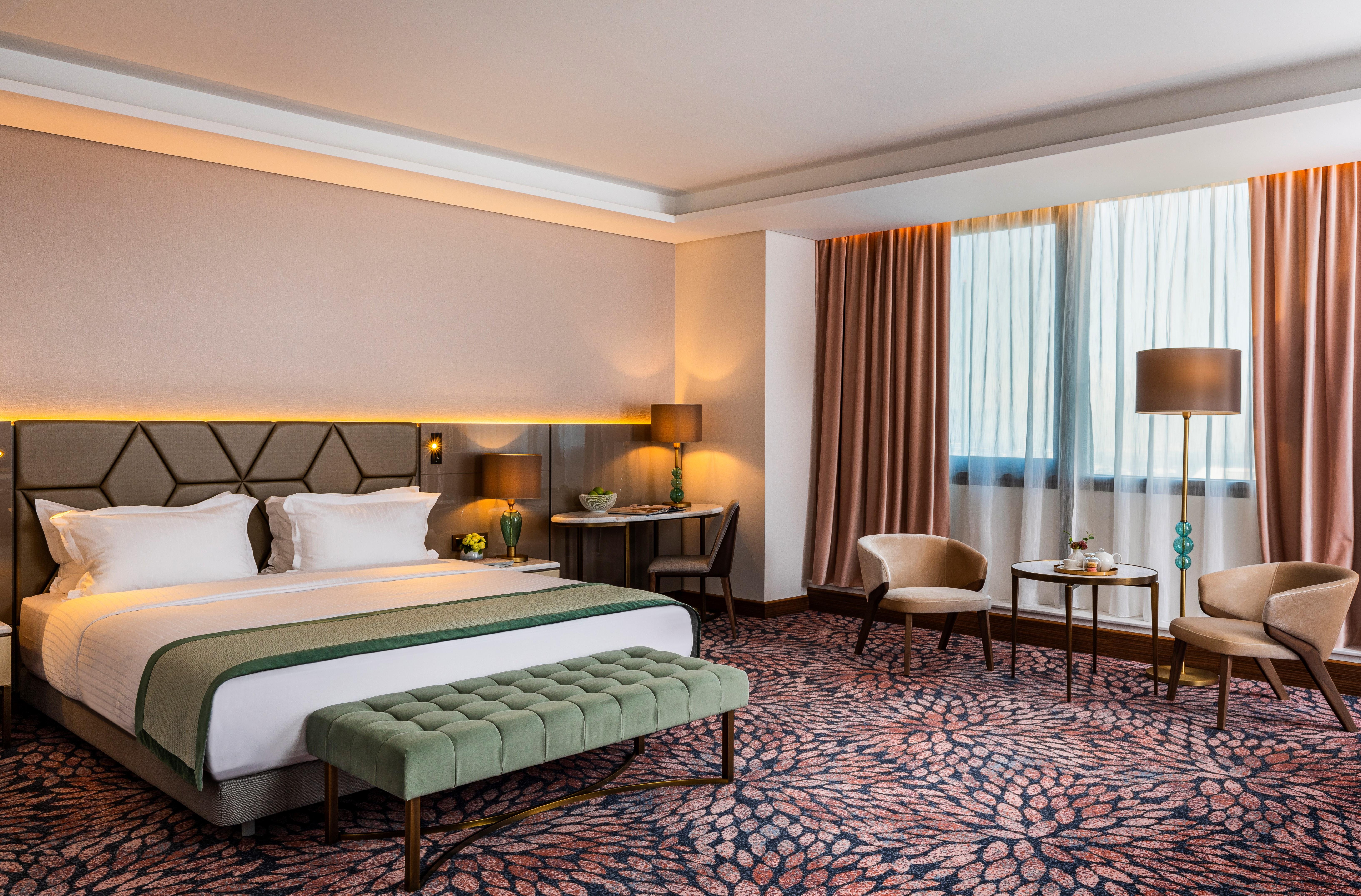 Grand Hotel Millennium صوفيا المظهر الخارجي الصورة The photo shows a modern hotel room. It features a large bed with a headboard, topped with two white pillows and a green throw blanket. In front of the bed, there is a stylish green bench. On either side of the bed, there are bedside tables with lamp