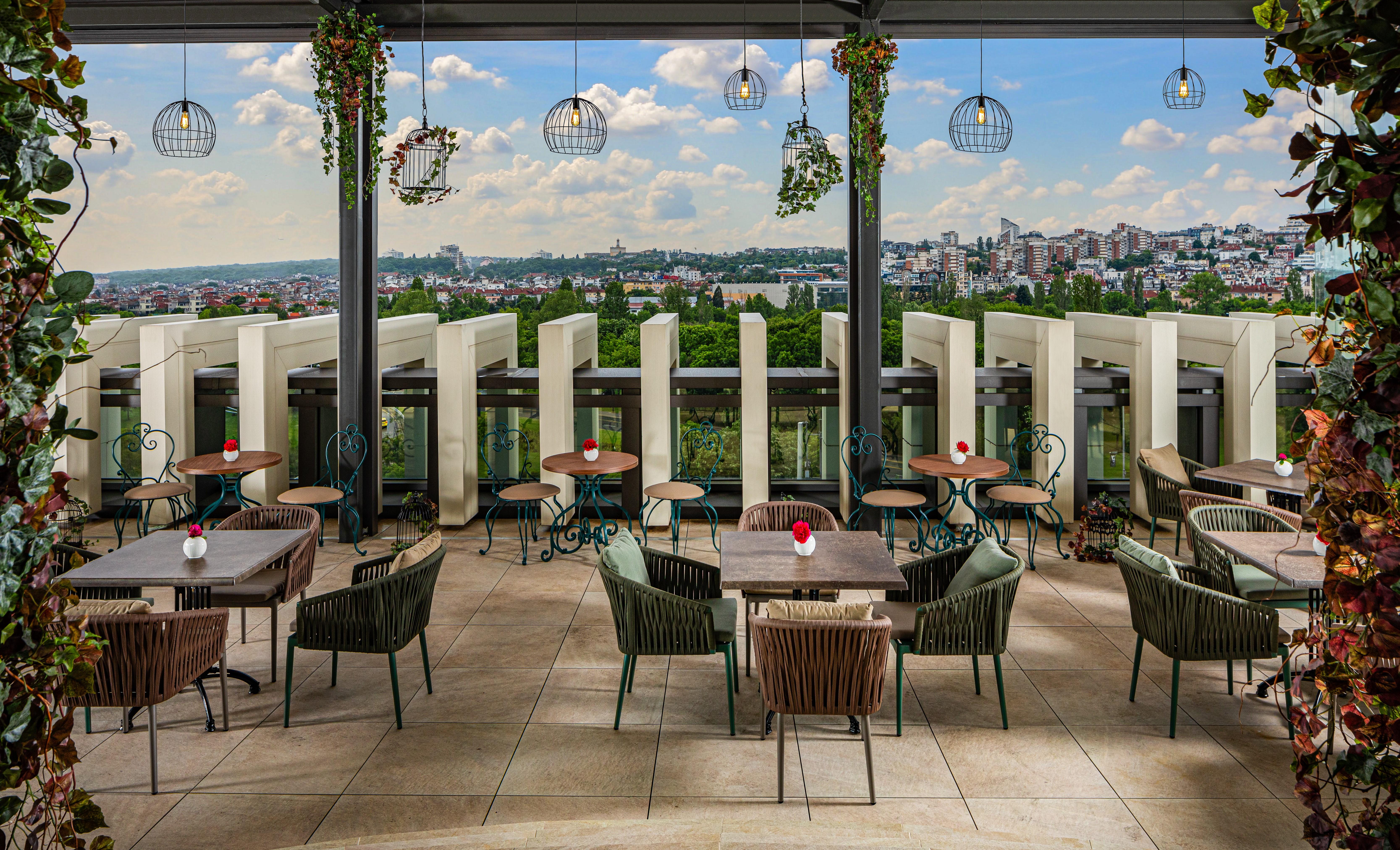 Grand Hotel Millennium صوفيا المظهر الخارجي الصورة The photo shows a stylish outdoor restaurant or café setting. There are several tables arranged on a tiled patio, each with chairs, some made of woven materials. Centerpieces with small red flowers adorn each table. The space is partially shaded by o