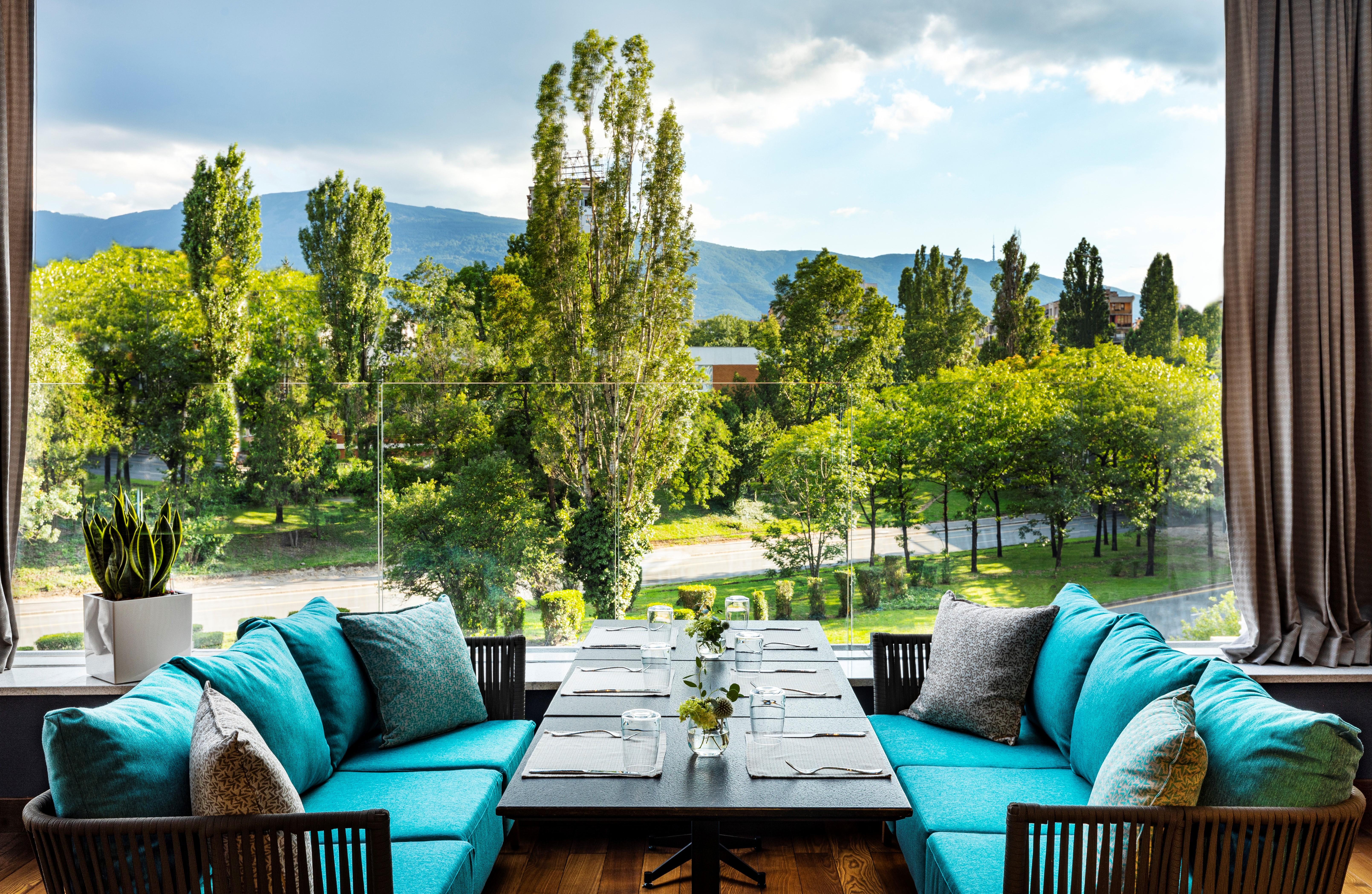 Grand Hotel Millennium صوفيا المظهر الخارجي الصورة The photo shows a bright and inviting dining area with large windows that overlook a lush green landscape. There are several trees in the background, along with a view of distant mountains under a partly cloudy sky. The seating consists of stylish tu