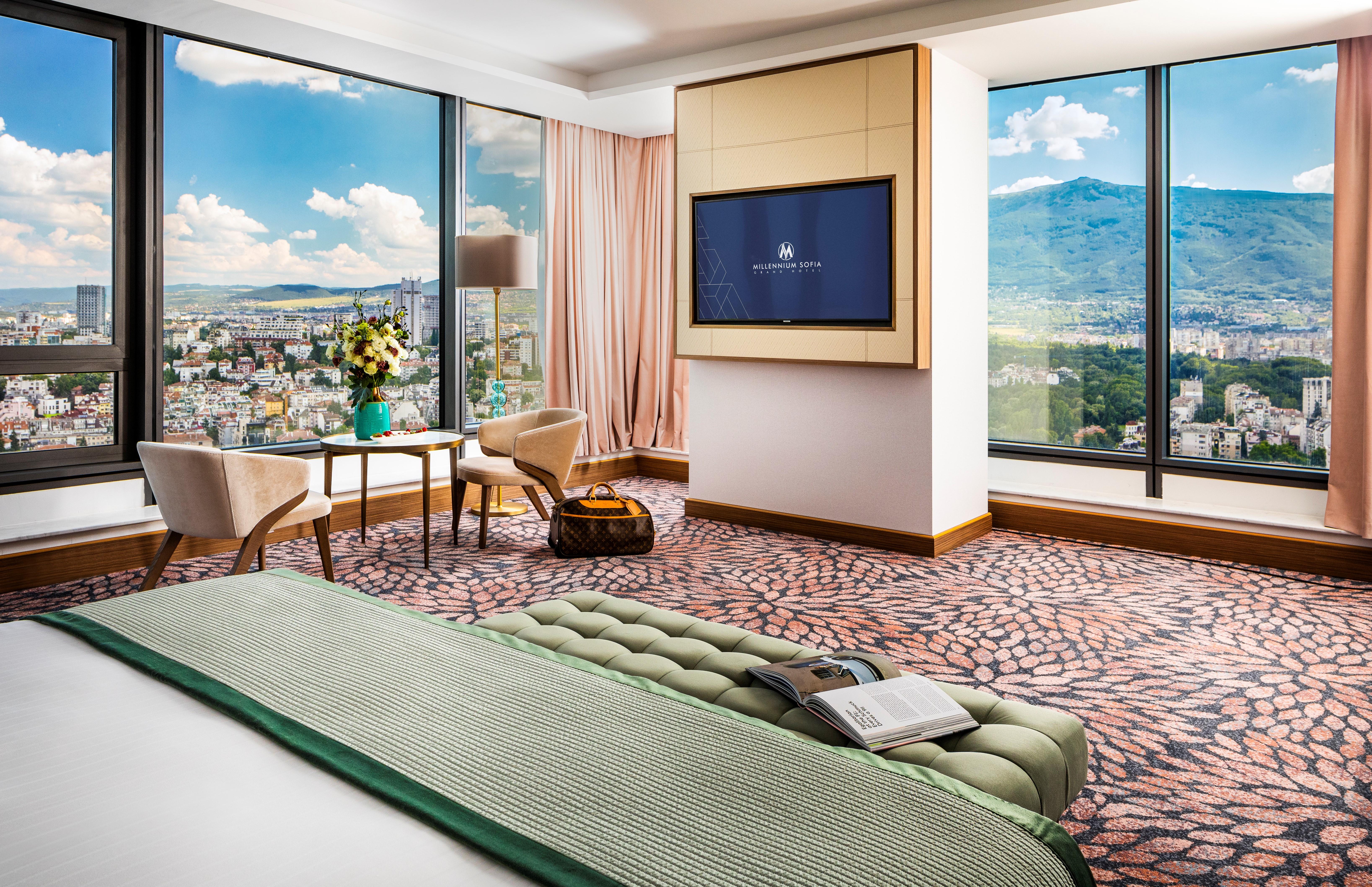 Grand Hotel Millennium صوفيا المظهر الخارجي الصورة The photo showcases a stylish hotel room with modern decor. It features large windows providing a panoramic view of a cityscape and mountains in the background. Inside, there is a comfortable bed with a green bedspread, a seating area with two armcha