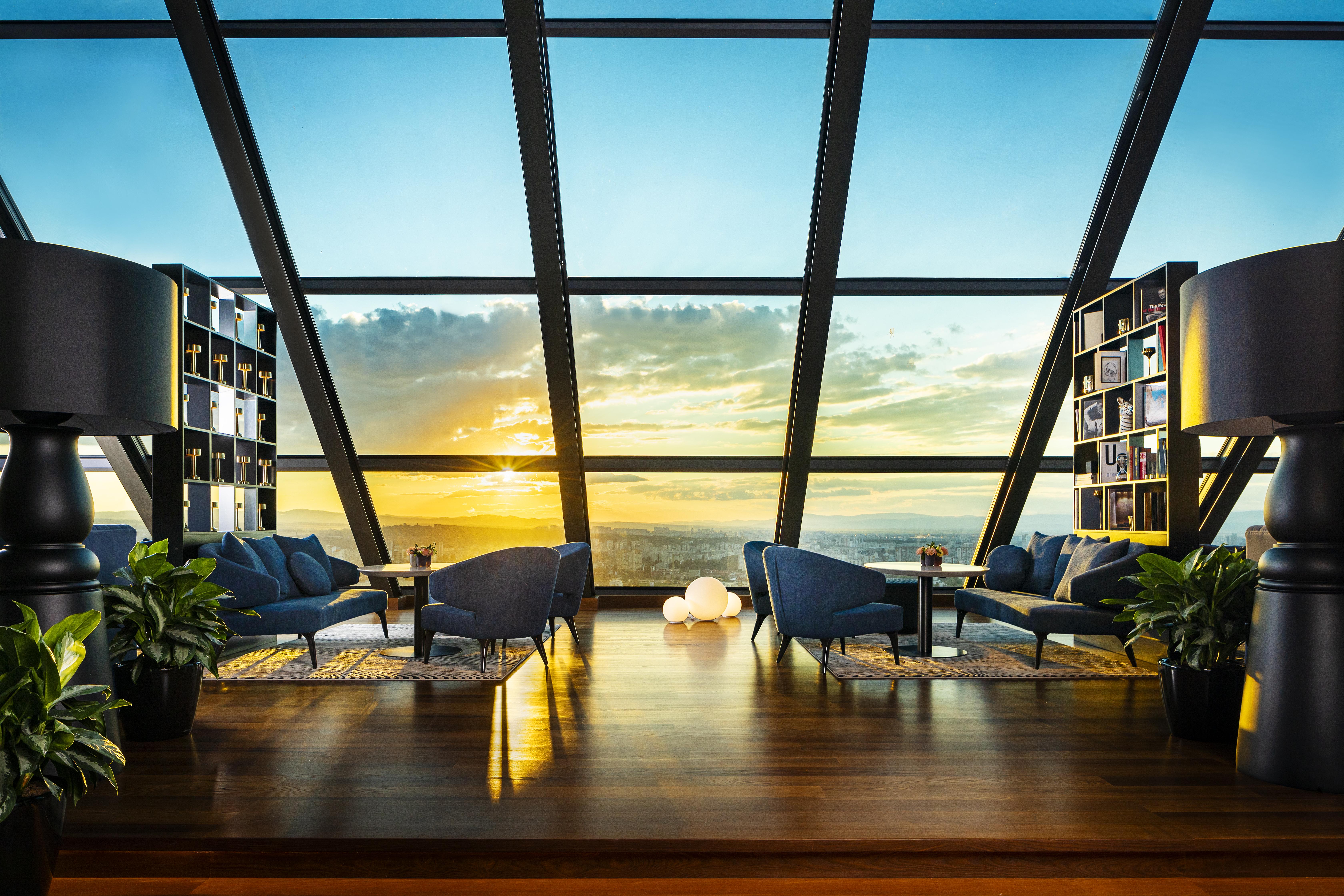 Grand Hotel Millennium صوفيا المظهر الخارجي الصورة The photo depicts a modern, stylish interior of a high-rise space with a panoramic view through large glass windows. The scene features a cozy seating area with several blue armchairs arranged around a low table, creating a comfortable atmosphere. Th