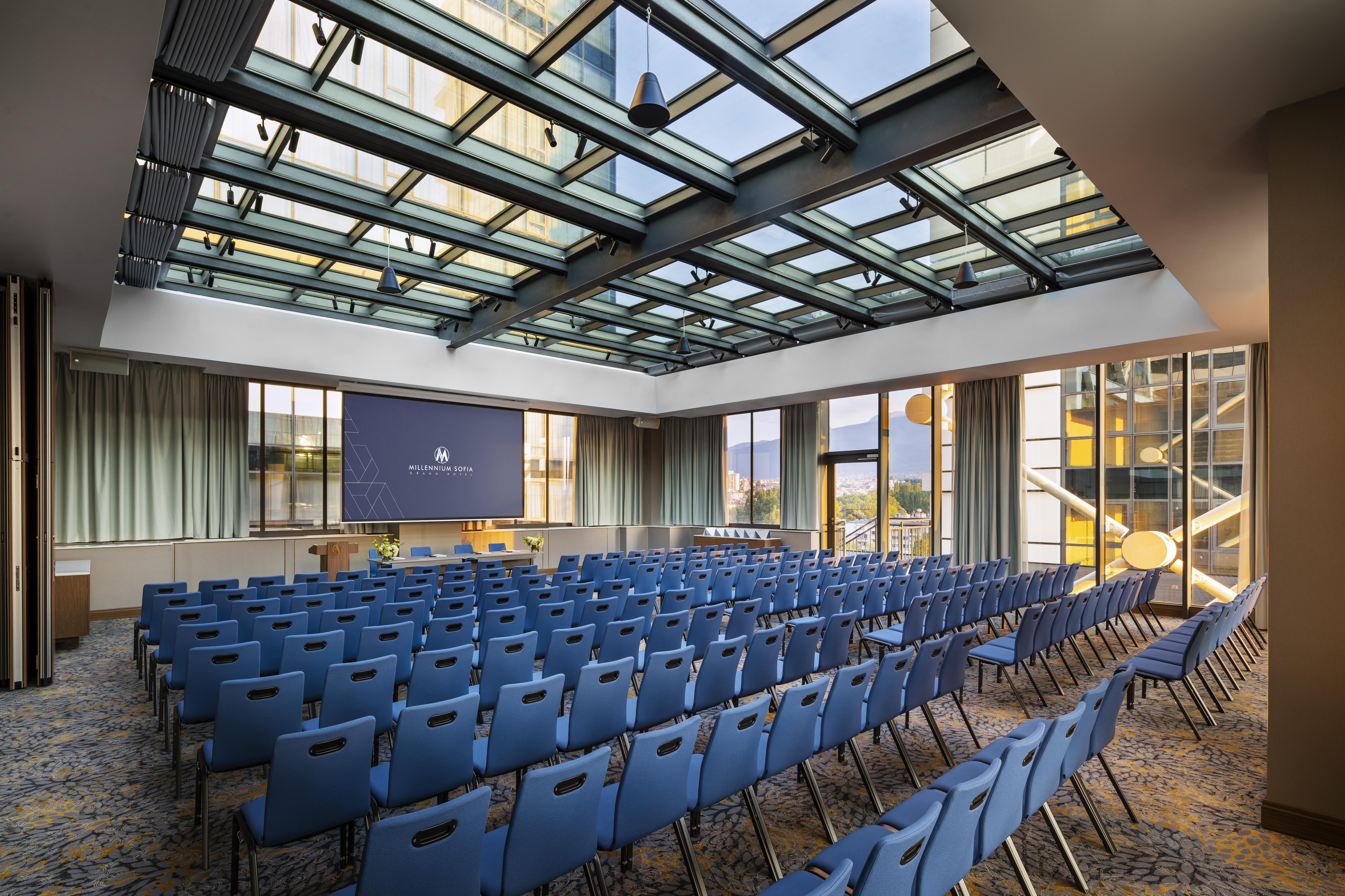 Grand Hotel Millennium صوفيا المظهر الخارجي الصورة The photo shows a large conference room or meeting space. It features rows of blue chairs arranged in a theater style, facing a large screen at the front of the room. The room is well-lit, with a glass ceiling allowing natural light to filter in. The