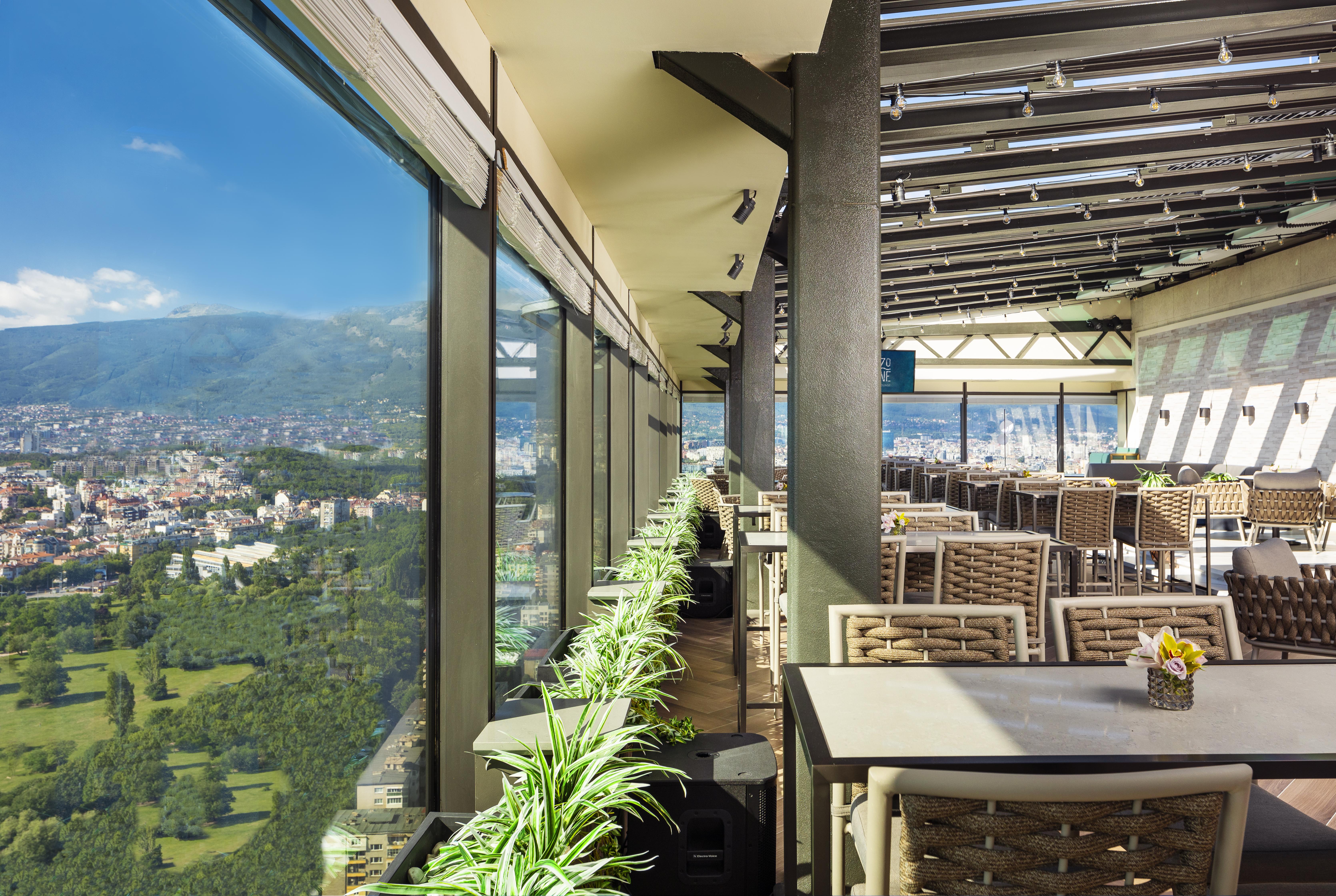 Grand Hotel Millennium صوفيا المظهر الخارجي الصورة The photo shows a modern, spacious restaurant with large glass windows offering a panoramic view of a city and surrounding landscape. The interior features contemporary furniture, including stylish tables and chairs. There are potted plants along the